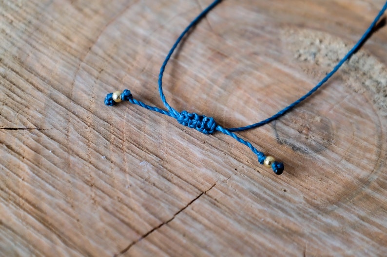 May include: A close-up of a blue adjustable bracelet with gold-colored beads. The bracelet is knotted in the center and rests on a textured wooden surface.
