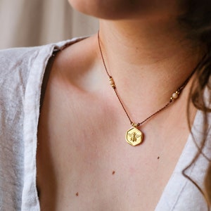 May include: A gold-colored pendant necklace with a bee design. The pendant hangs from a brown cord with small gold beads and light-colored stones. The necklace is worn against a person's skin, with a white shirt visible.