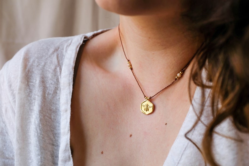 May include: A gold-colored pendant necklace with a bee design. The pendant hangs from a brown cord with small gold beads and light-colored stones. The necklace is worn against a person's skin, with a white shirt visible.
