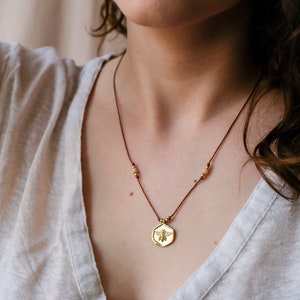 May include: A gold-colored pendant necklace featuring a bee design. The pendant hangs from a brown cord necklace with small, gold-colored beads. The necklace is worn over a light-colored top.