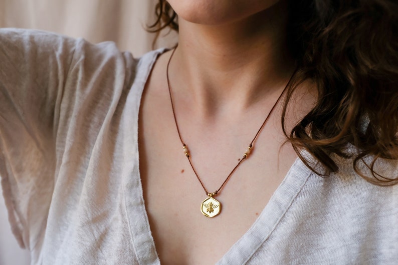May include: A gold-colored pendant necklace featuring a bee design. The pendant hangs from a brown cord necklace with small, gold-colored beads. The necklace is worn over a light-colored top.