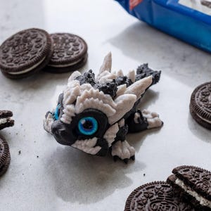 May include: A small, articulated dragon figurine featuring a black and white textured body with bright blue eyes, positioned among several round chocolate sandwich cookies with white cream filling and a blue package labeled Oreo.