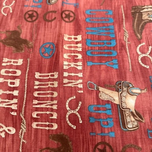 Pode incluir: Tecido vermelho com tema Western. O design inclui imagens de botas de cowboy, selas, cavalos, ferraduras e estrelas. As palavras "Cowboy Up", "Buckin' Bronco" e "Ropin' & Ridin'" são impressas em branco e azul.
