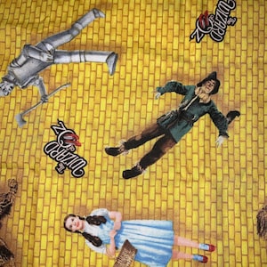 Può includere: Un tessuto giallo con personaggi de Il Mago di Oz. L'Uomo di latta, lo Spaventapasseri, Dorothy e il Leone Codardo sono raffigurati su uno sfondo di strada di mattoni gialli. Sono presenti anche le parole "The Wizard of Oz".