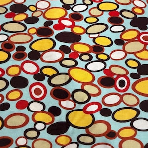 Könnte beinhalten: Stoff mit einem sich wiederholenden Muster aus farbigen Ovalen in Gelb, Braun, Rot und Weiß auf hellblauem Hintergrund. Das Design erinnert an einen Retro- oder Mid-Century-Modern-Stil.
