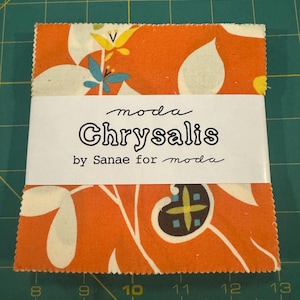 Puede incluir: Un cuadrado de tela con fondo blanco y un patrón floral naranja. La tela está etiquetada con "moda Chrysalis by Sanae for moda".