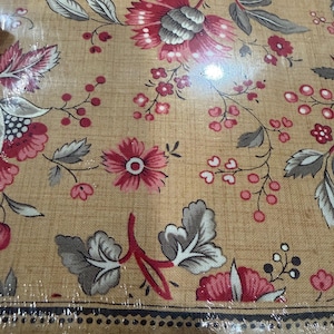 May include: Close-up of a floral fabric with a tan background. The fabric features large, detailed flowers in shades of red and pink, with grey and white leaves. Small red berries and delicate floral accents are scattered throughout the design. The fabric has a decorative border.