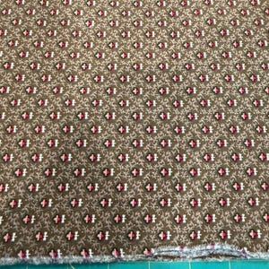 May include: Brown fabric with a repeating pattern of small, stylized floral shapes in red, black, and white. The fabric has a slightly textured appearance and a raw edge along the bottom.