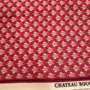 May include: Red fabric featuring a repeating white and grey floral design. The fabric has a border with the text "CHATEAU ROUGE" printed on it. The material is likely cotton or a similar fabric, ideal for quilting or sewing projects.