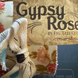 Può includere: Un primo piano di un'etichetta in tessuto con il testo "Gypsy Rose" e "By Fig Tree Quilts" in uno schema di colori marrone, crema e verde. L'etichetta è attaccata a un tessuto bianco con un motivo floreale.