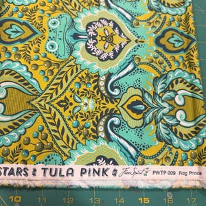 May include: Close-up of a fabric with a repeating paisley design in shades of yellow, teal, and navy. The fabric has the text "STARS TULA PINK" and "Free Spirit" printed on it. The fabric is on a green cutting mat.