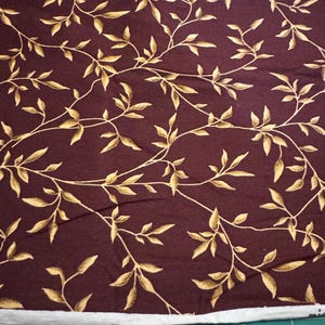 May include: A burgundy fabric with a repeating pattern of gold leaves and branches.