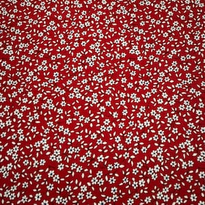 May include: A close-up of a red fabric with a repeating pattern of small white flowers and green leaves. The floral design is densely packed across the entire surface, creating a textured appearance.