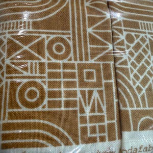 May include: Close-up of fabric with a geometric design in white on a tan background. The pattern includes circles, squares, and lines. The text "The secret of happiness is freedom..." is printed at the bottom.