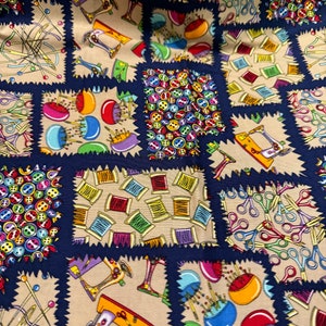 Könnte beinhalten: Ein beigefarbener Stoff mit einem Patchwork-Muster mit verschiedenen Nähmuster. Die Muster umfassen Knöpfe, Garnrollen, Nähmaschinen und Scheren. Der Stoff ist mit einem dunkelblauen, gewellten Rand eingefasst.