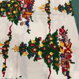 Pode incluir: Tecido branco com um padrão repetitivo de árvores de Natal. As árvores são adornadas com enfeites, presentes e uma estrela. Laços vermelhos e sinos acentuam o design. O texto "For Alexander Henry" está impresso na parte inferior.