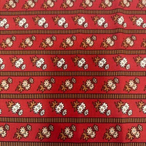 May include: Red fabric with a repeating floral pattern. The design features clusters of white and tan flowers with brown leaves, set against a red background. Horizontal stripes with brown and black lines separate the floral arrangements.