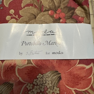 以下が含まれることがあります： 赤、茶色、クリーム色の花柄の生地見本です。生地には「moda Portobello Market by 3 Sisters for moda」とラベルが貼られています。