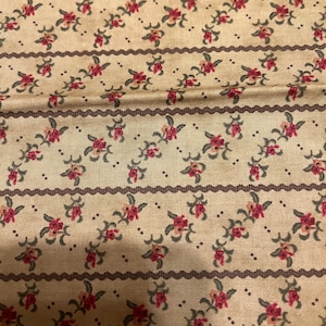 Op de afbeelding: Close-up van een beige stof met een herhalend bloemenpatroon. Het ontwerp toont kleine rode bloemen met groene bladeren, gerangschikt in diagonale rijen. Donkerbruine golvende lijnen en stippen scheiden de bloemenrijen.
