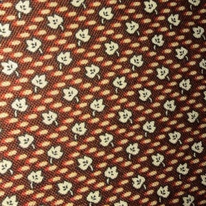 May include: Close-up of a fabric featuring a repeating pattern of white floral shapes on a dark brown background. The design includes diagonal rows of small, light brown ovals and red dashes.