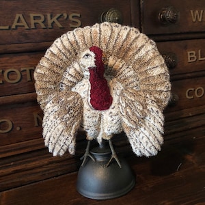 May include: A brown and white turkey with a red head and neck, standing on a black metal base. The turkey is made of fabric and has a detailed design.