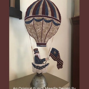 May include: A punch needle design of a hot air balloon with a figure holding a flag. The balloon is red, white, and blue with a beige pattern. The figure is wearing a blue coat and a red, white, and blue hat. The flag is red, white, and blue with a star in the upper left corner. The design is titled "Freedoms Flight" and is an original design by Colleen Colaizzi. Copyright Colleen Colaizzi 2021.