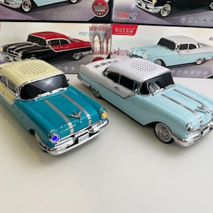 Puede incluir: Dos altavoces con forma de coche de juguete en color turquesa y azul claro, con detalles cromados. Los altavoces están diseñados para parecerse a coches clásicos, con el texto "WSTER" visible en la parte delantera. El embalaje presenta imágenes de altavoces de coche similares.