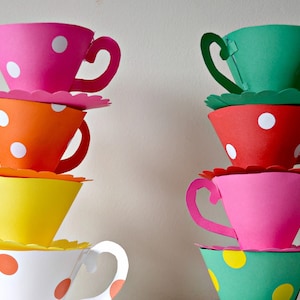 Puede incluir: Una pila de cuatro tazas de té de papel de colores con lunares blancos. Las tazas están apiladas en forma de pirámide, con la taza más grande en la parte inferior y la taza más pequeña en la parte superior. Las tazas están hechas de papel y tienen un borde festoneado.