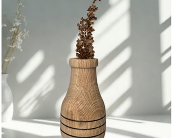 May include: A wooden vase with a light brown finish and three dark bands around the base. The vase holds a dried, brown floral arrangement. The vase is set against a white background with shadows.