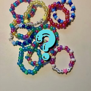 May include: A collection of colorful beaded bracelets with letter beads spelling out words like "SUNDROP", "RAWR", and "XD".