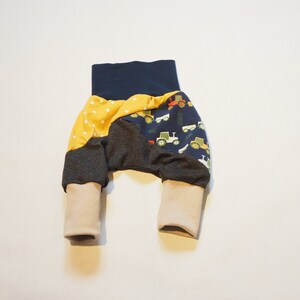Può includere: Un paio di pantaloni per neonati con un design a patchwork. I pantaloni sono realizzati in un tessuto morbido ed elastico e hanno uno schema di colori blu navy, grigio e giallo. I pantaloni presentano una stampa di trattori sulle gambe e pois bianchi sul tessuto giallo.