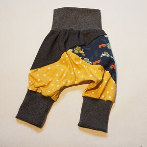 May include: Baby trousers with a grey waistband and cuffs. The trousers feature a patchwork design with a mustard yellow polka dot pattern, navy blue fabric with tractor graphics, and grey fabric.