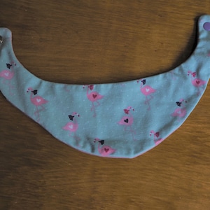 May include: A light blue baby bib with a pink flamingo pattern. The bib has two snaps on the sides.