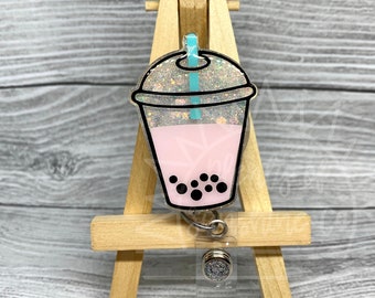 May include: A pink and white glitter bubble tea charm with a black and white bubble design. The charm is attached to a silver retractable badge reel with a clear plastic case.
