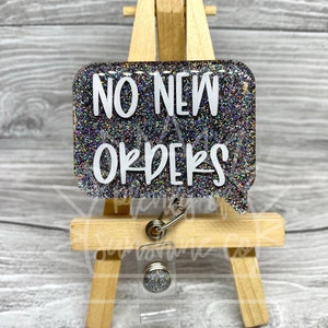 May include: A silver glitter badge holder with a speech bubble that says "NO NEW ORDERS".