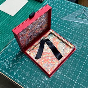 May include: Open red box with a patterned interior and a black ribbon. The box is on a green cutting mat with a grid pattern. The interior of the box has a colorful, marbled design. The box is open, revealing the inside.