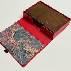 May include: A vintage book in a red box. The book has a worn, brown leather cover. The box is open, revealing a marbled paper interior with grey, red, and yellow patterns. A black ribbon is attached to the box.