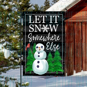 May include: A garden flag with the text "LET IT SNOW Somewhere Else" and a snowman illustration. The snowman has a carrot nose and holds a red bird. Green pine trees are behind the snowman, and the background is black and white.