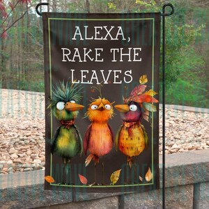 May include: A garden flag with the text "ALEXA, RAKE THE LEAVES" and three cartoon birds. The birds are in shades of green, orange, and yellow, with fall leaves. The flag is rectangular and has a black background.