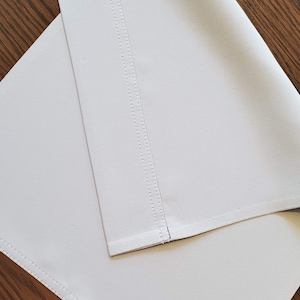 May include: Two folded, light gray fabric pieces with stitched edges and seams. The fabric has a subtle texture and appears to be a durable material. The pieces are arranged at an angle against a wooden surface.