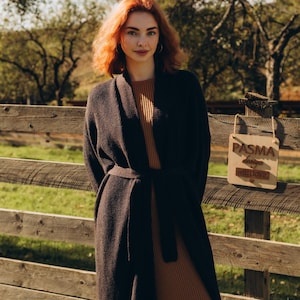 Può includere: Una donna con i capelli rossi indossa un cardigan marrone e un vestito marrone e si trova davanti a una recinzione di legno. Un cartello di legno con il testo "PASMA design with love" è appeso alla recinzione.