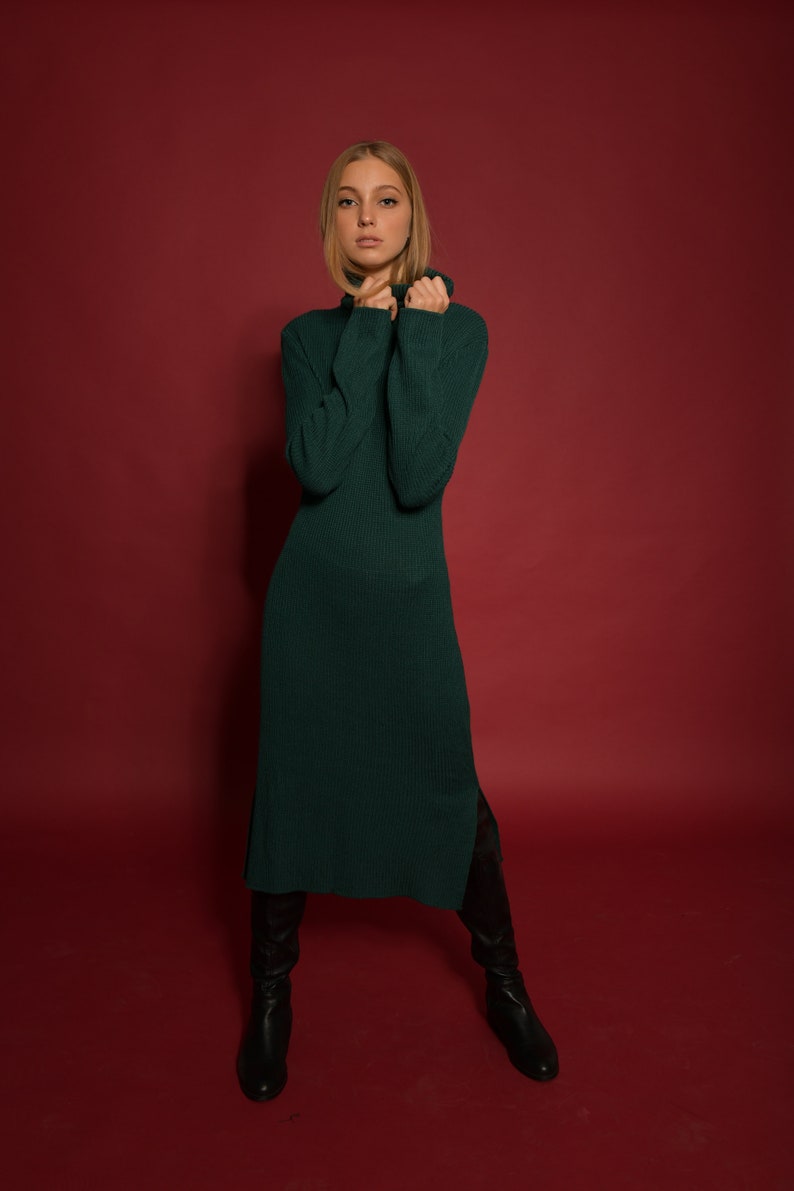 May include: A woman wearing a long, dark green turtleneck sweater dress with a side slit. She is standing on a red background and wearing black knee-high boots.