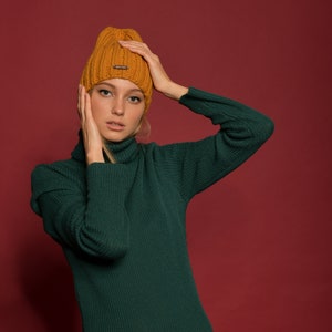 May include: A woman wearing a green turtleneck sweater and a mustard yellow knit beanie. The beanie has a small brown logo on the front.
