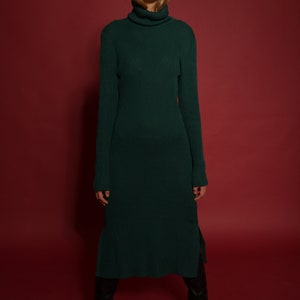 May include: A woman wearing a long, dark green turtleneck sweater dress with a ribbed texture. She is standing on a red background and wearing black knee-high boots.