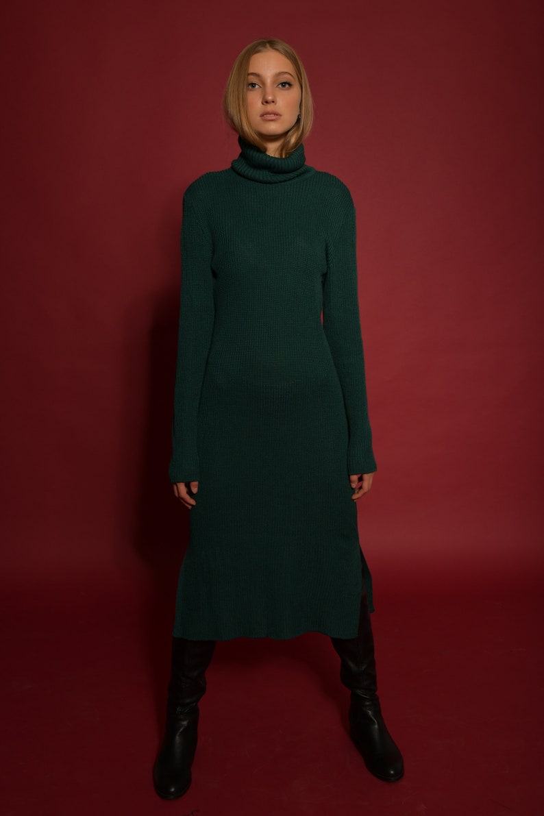 May include: A woman wearing a long, dark green turtleneck sweater dress with a ribbed texture. She is standing on a red background and wearing black knee-high boots.
