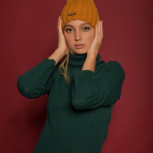 May include: A woman wearing a green turtleneck sweater and a mustard yellow knit beanie. She is posing with her hands on her cheeks.