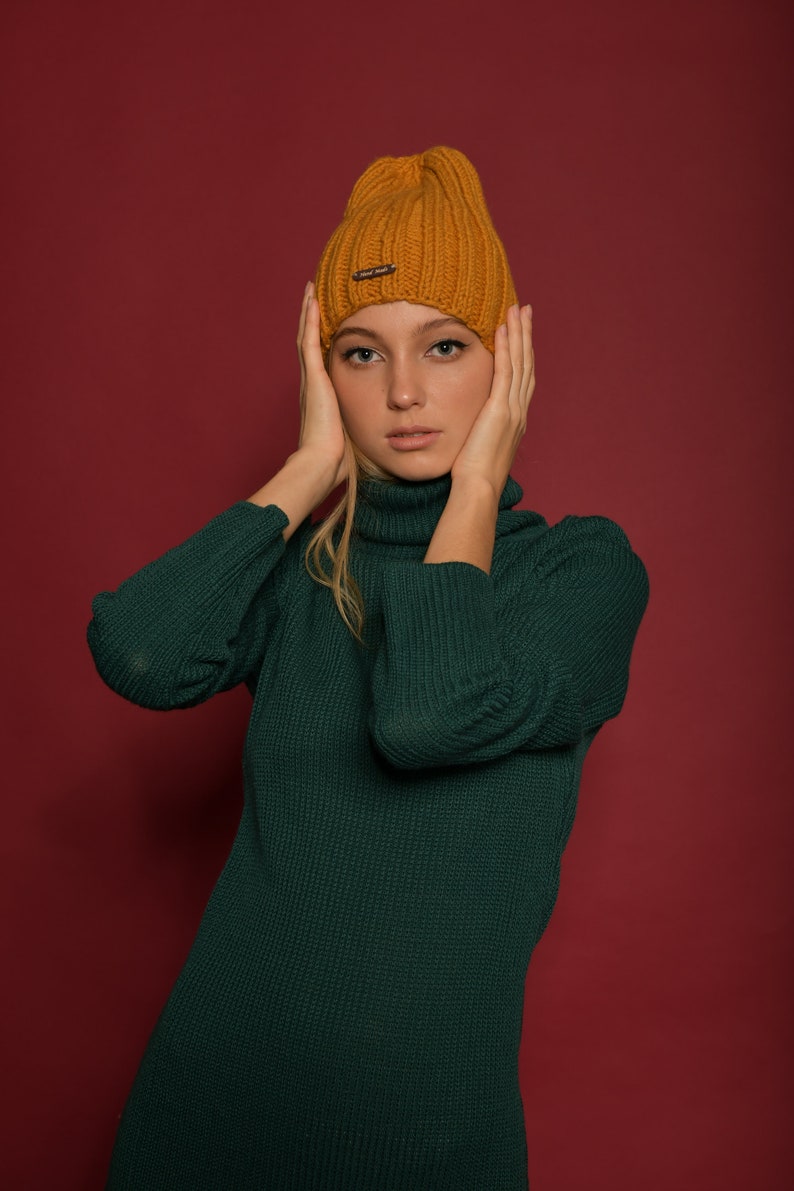 May include: A woman wearing a green turtleneck sweater and a mustard yellow knit beanie. She is posing with her hands on her cheeks.