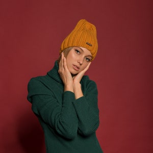 May include: A woman wearing a green turtleneck sweater and a mustard yellow knitted beanie. She is posing with her hands on her cheeks.