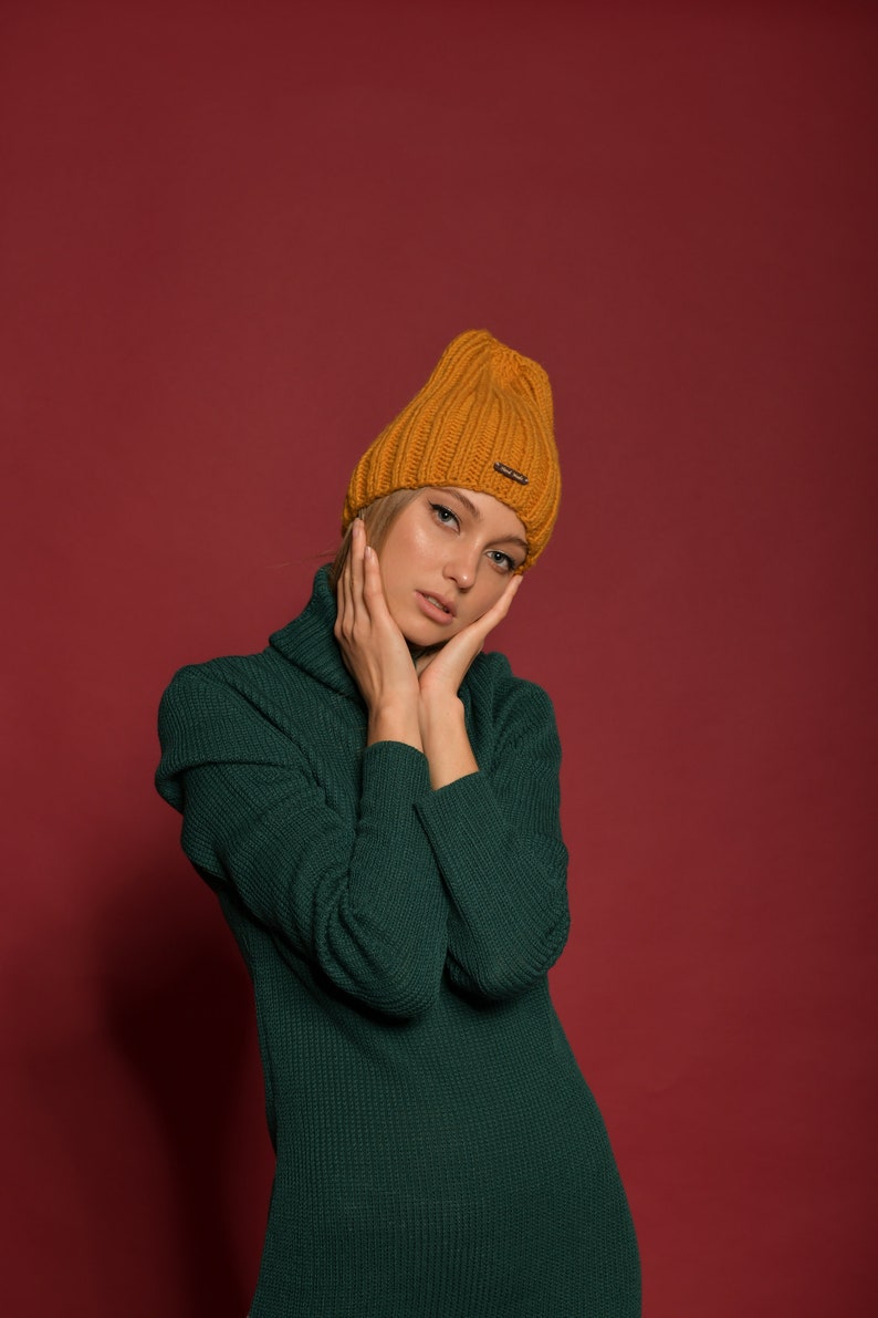 May include: A woman wearing a green turtleneck sweater and a mustard yellow knitted beanie. She is posing with her hands on her cheeks.