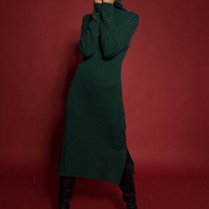 May include: A woman wearing a long, dark green turtleneck sweater dress with a side slit. She is wearing black knee-high boots and standing on a red background.